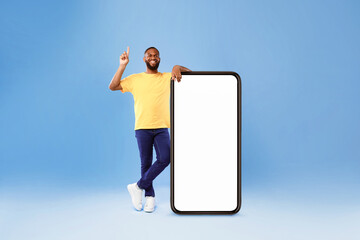 Poster - Handsome millennial African American guy offering mockup for new mobile app on giant smartphone screen, pointing upwards on blue studio background. Space for online advertisement design