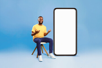 Poster - Attractive African American man sitting on chair next to huge smartphone with blank white screen, advertising new mobile application on blue studio background, mockup for website or ad design