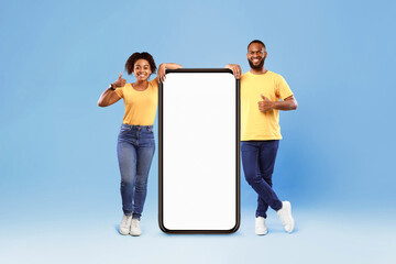Poster - Cool mobile app. Young African American couple standing near giant smartphone with mockup, showing thumbs up, promoting application or website on blue studio background, full length portrait