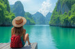 © INVO - Traveler sitting on a wooden dock, wearing a straw hat and red backpack, admiring the stunning green limestone mountains of Phang Nga Bay. A serene and picturesque travel scene capturing adventure,