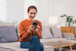 © kenchiro168 - Woman in cozy orange sweater sits on sofa, smiling while using her smartphone in bright living room.