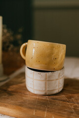 Two yellow and white ceramic cups are stacked on top of each other on a wooden cutting board. The cups have a checkered design and appear to be handmade. Concept of warmth and comfort