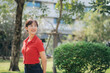 © Jirawatfoto - Asian cheerful woman in red shirt smiles while standing in sunny park surrounded by greenery and trees