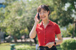 © Jirawatfoto - Asian cheerful woman in red shirt talking on phone while standing outdoors in sunny park