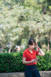 © Jirawatfoto - Asian young woman in red shirt and jeans stands outdoors, holding smartphone while surrounded by lush greenery and sunlight