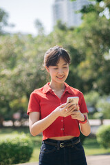 Wall Mural - Asian young woman in red shirt smiles while using her smartphone outdoors in sunny park, surrounded by greenery and trees