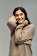 © LIGHTFIELD STUDIOS - Young plus size woman smiles warmly in a beige sweater against a grey backdrop