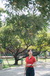 © Jirawatfoto - Asian woman in red shirt and jeans walks through sunny park, surrounded by lush green trees and serene atmosphere