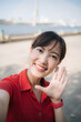 © Jirawatfoto - Asian cheerful woman in red shirt smiles and waves while taking selfie outdoors near river on sunny day