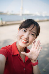 Wall Mural - Asian cheerful woman in red shirt smiles and waves while taking selfie outdoors near river on sunny day