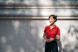 © Jirawatfoto - Asian woman in red shirt stands outdoors against white wall, smiling warmly under dappled sunlight