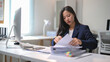 © Wasana - A woman is sitting at a desk with a computer monitor and a stack of papers