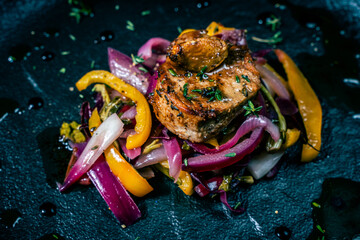 Wall Mural - Slice of roasted pork tenderloin with vegetables, pink onion and yellow pepper on a black plate on a dark background. Beautiful, restaurant presentation of the dish.