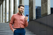 © Liubomir - Successful man walking through the city outside an office building with a phone in his hands. Businessman using an app on his smartphone, typing messages, and browsing social media, smiling.