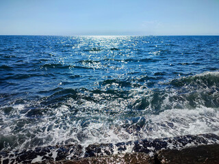 Beautiful clean sea on sunny warm day. Surface of blue calm sea with sun glare and blue sky. Sea waves with white foam crashing against old stones on shore. Nature seascape, sea landscape. Vertical