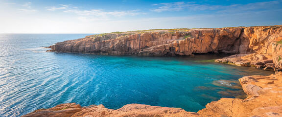 Poster - A panoramic photo of a sunlit rocky coastline with turquoise waters, showcasing the rugged natural beauty of a secluded bay and the peaceful ocean under a clear sky.