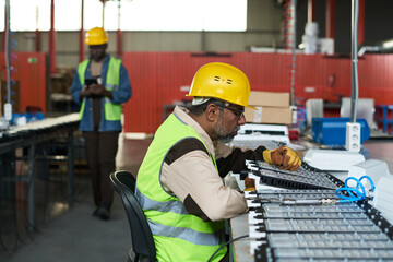 Leinwandbilder - Multiethnic mature engineer using electric handtool in workshop against young colleague holding mobile gadget