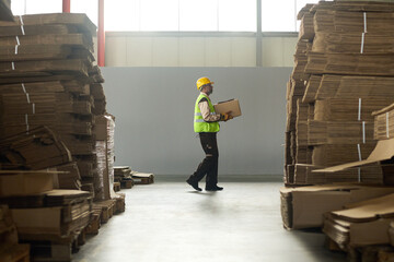 Leinwandbilder - Long shot of male engineer carrying packed box on hands while moving between tall stacks of cardboard sheets in workshop