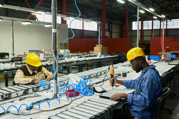 Aufkleber - Intercultural engineers in hardhats using electric equipment while sitting by long tables and producing new spare parts