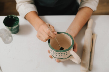 Poster - Female artist is painting earthenware with paintbrush and water colors in the workshop. Painter is drawing and making a colorful decoration of a clay ware.