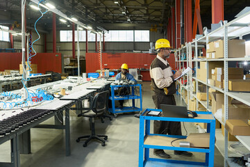 Leinwandbilder - Serious mature worker of factory standing in front of shelves with packed boxes and taking notes in document during revision