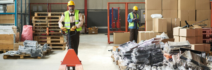 Leinwandbilder - Header of mature engineer in workwear pushing empty pallet truck while moving along stacks of packed items in warehouse or plant