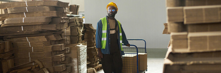 Poster - Header of young warehouse worker in workwear pulling cart with packed boxes while moving between stacked cardboard sheets