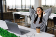 © F8  \ Suport Ukraine - Young business woman smiling and relaxing in a bright office, enjoying a break at work with hands behind her head.