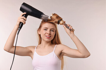 Wall Mural - Beautiful young woman styling her hair with hairdryer and brush on light grey background