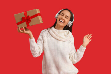 Wall Mural - Happy young woman in headphones with Christmas gift box on red background