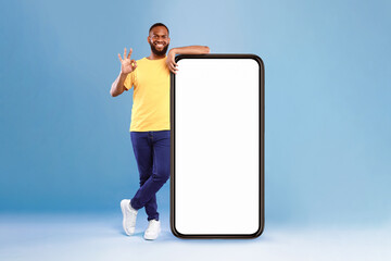 Poster - Positive African American guy showing okay gesture next to huge smartphone on blue studio background, empty space for mobile app design on screen. Website or advertisement mockup