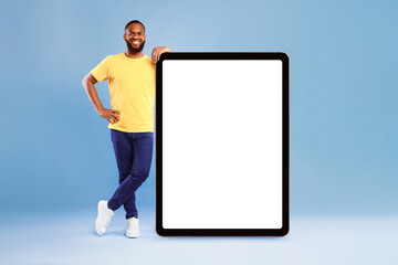 Poster - Full length portrait of cheerful young African American guy leaning on giant tablet pc, demonstrating blank screen on blue studio background, free space for online advertisement, mockup for newest app