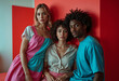 © The - Three young adults, a blonde woman in a pink and teal dress, a brunette woman in a beige shirt and floral pants, and a Black man in a teal shirt, pose closely together against a red wall. Their fashio