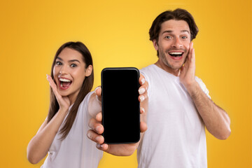 Poster - Advertising New Mobile App. Portrait of happy excited couple holding smartphone with black empty screen in hands, showing device with blank space for mock up, saying wow, orange studio background