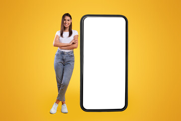 Aufkleber - Full body length of smiling casual lady standing with folded arms near giant mobile phone, empty white screen template, orange studio background. Mock up for app or website, free space for advertising