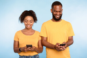 Poster - Positive young African American couple using cellphones, surfing web, posting in social media, checking email or sms messages on blue studio background. Online life, modern gadgets concept