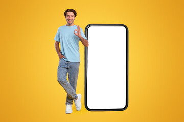 Poster - Cool Mobile Offer. Happy casual guy standing near big huge cellphone with empty white screen, showing ok, recommending great new app or website for cell phone, orange wall, mock up, full body length