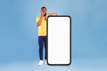 Poster - Shocking mobile offer. Emotional African American guy leaning on giant smartphone with blank screen, biting nails in panic, afraid to miss huge web store sale on blue background, mockup