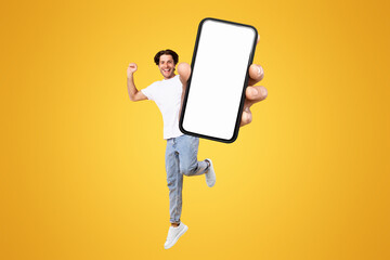 Poster - Yes, Cool Mobile Offer Ad. Joyful young casual guy showing big empty cell phone screen jumping up in the air on orange studio background, creative collage. Happy man recommend website, free copy space