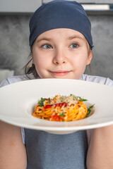 Wall Mural - Portrait of a beautiful little girl in a gray bandana and apron holding a plate of pasta in her hands. The girl shows the spaghetti she has prepared. Close-up.