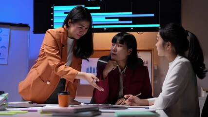 Wall Mural - Asian businesswomen working late, analyzing data, and collaborating on a project in a brightly lit office, demonstrating teamwork and dedication