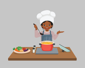 Wall Mural - Cute little African girl chef cooking delicious vegetable soup with hand showing for copy space
