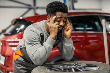 Fototapete - Multiracial car service worker leaning on pile of tires at mechanic workshop and rubbing his head because of headache.