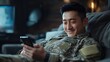 © Kritsanaphong - Connecting from Afar: A soldier in camouflage uniform relaxes on a couch, a warm smile lighting up his face as he uses his smartphone.