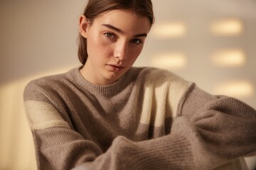 Wall Mural - A young woman with brown hair wearing a light brown and cream colored sweater looks directly at the camera.