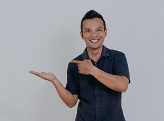 Wall Mural - Confident smiling asian man with left hand pointing to his opened right hand palm, isolated on white background.