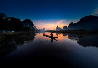 Poster - Serene China landscape at twilight. Calm river reflects the colorful sky and silhouette of a person in a boat surrounded by mountains.