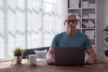 Wall Mural - Confident smiling senior man using laptop, working from home sitting at wooden table with coffee cup and plant, window and bookshelf in background