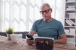 © MINAE - Mature man with eyeglasses using digital tablet and smartphone while working or relaxing at home, sitting at wooden table with a cup of coffee or tea
