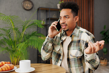 Canvas Print - Young sad mad man of African American ethnicity wear grey casual clothes talk speak on mobile cell phone sitting alone at table in coffee shop cafe relax rest in restaurant during free time indoors.